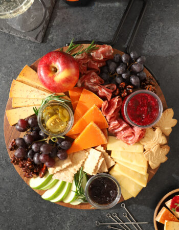 a round platter of assorted snacks, including slices of cheese crackers, grapes, candied walnuts, apples, a jar of red jam, salami and sausage, and maple leaf shaped cookies. There is a bottle of champagne off to the side next to a stack of small black plates.