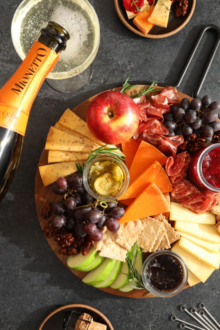 a round platter of assorted snacks, including slices of cheese crackers, grapes, candied walnuts, apples, a jar of red jam, salami and sausage, and maple leaf shaped cookies. There is a bottle of champagne off to the side next to a stack of small black plates.