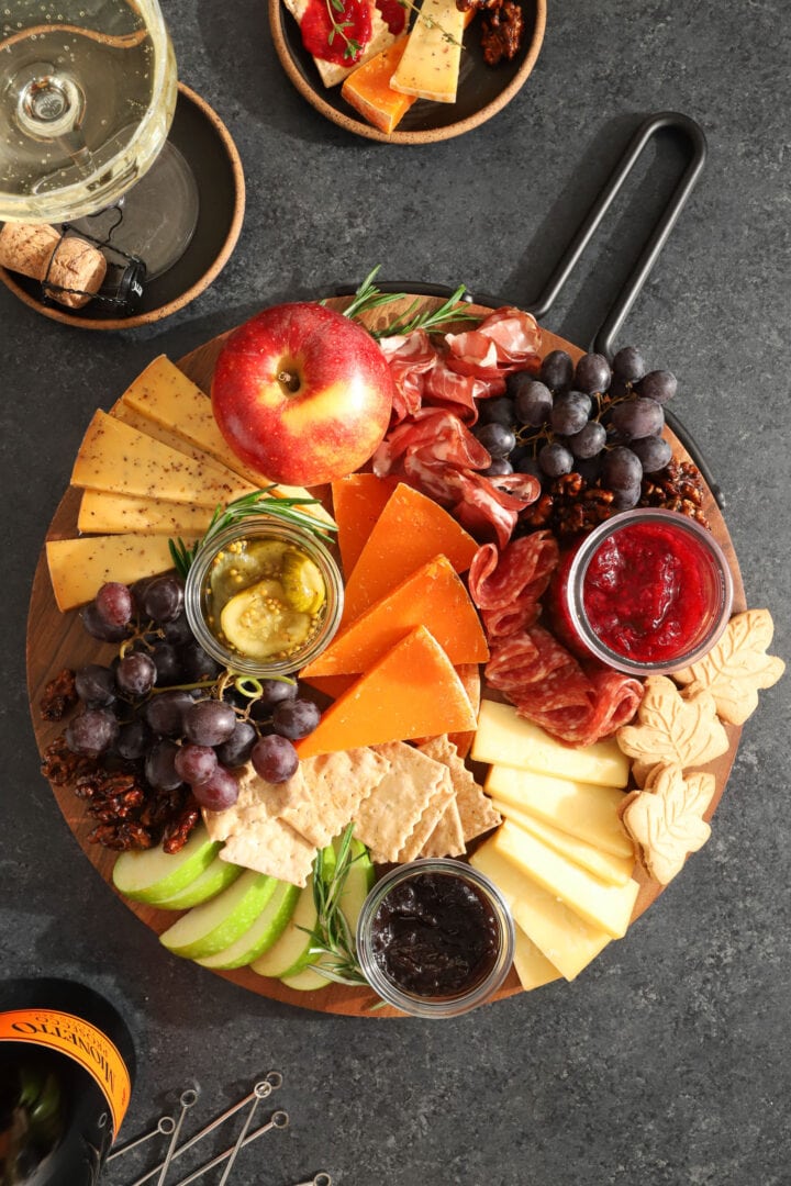 a round platter of assorted snacks, including slices of cheese crackers, grapes, candied walnuts, apples, a jar of red jam, salami and sausage, and maple leaf shaped cookies. There is a bottle of champagne off to the side next to a stack of small black plates.