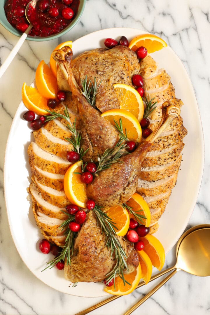 roasted and carved turkey on a white platter, garnished with fresh rosemary sprigs, cranberries and orange slices