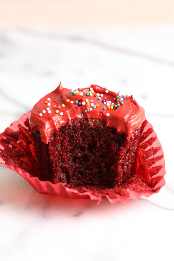 an unwrapped red velvet cupcake with red velvet frosting and rainbow sprinkles