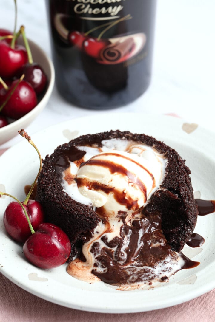 a small chocolate cake on a white plate topped with vanilla ice cream and garnished with fresh cherries.
