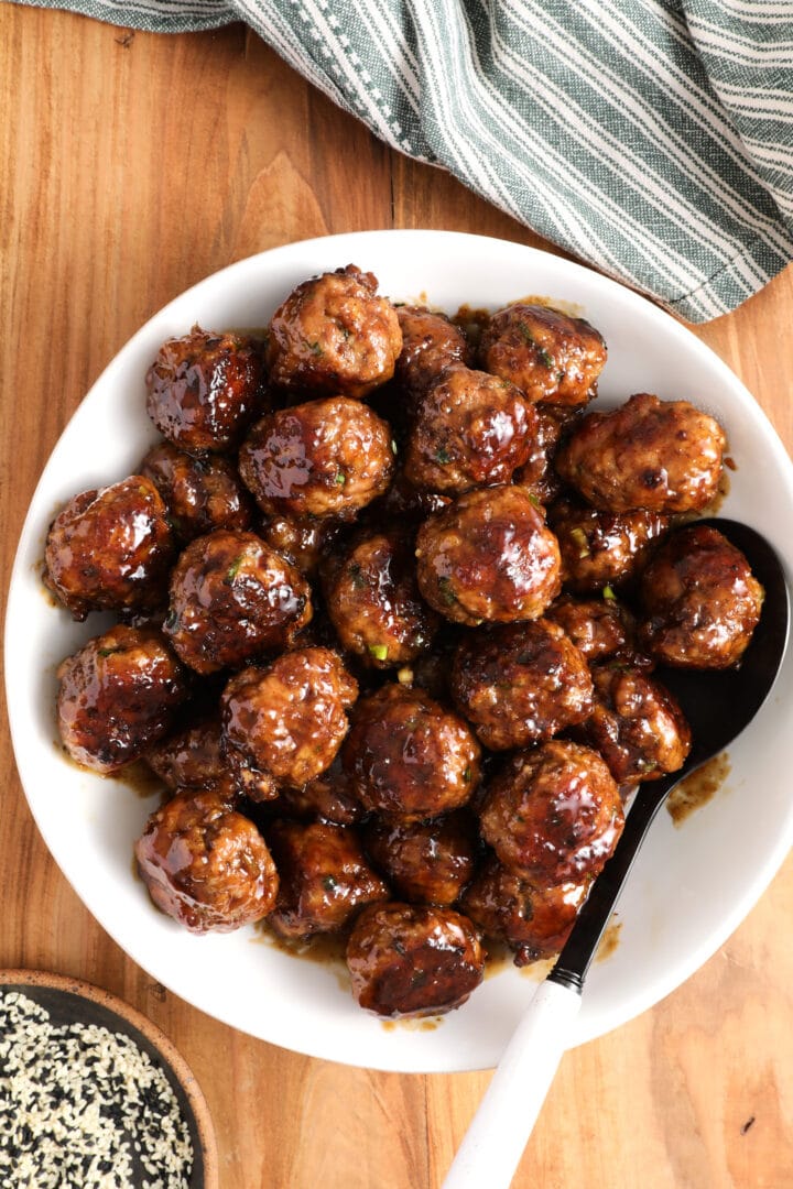 white bowl of teriyaki meatballs with a large spoon and bowl of sesame seeds off to the side.