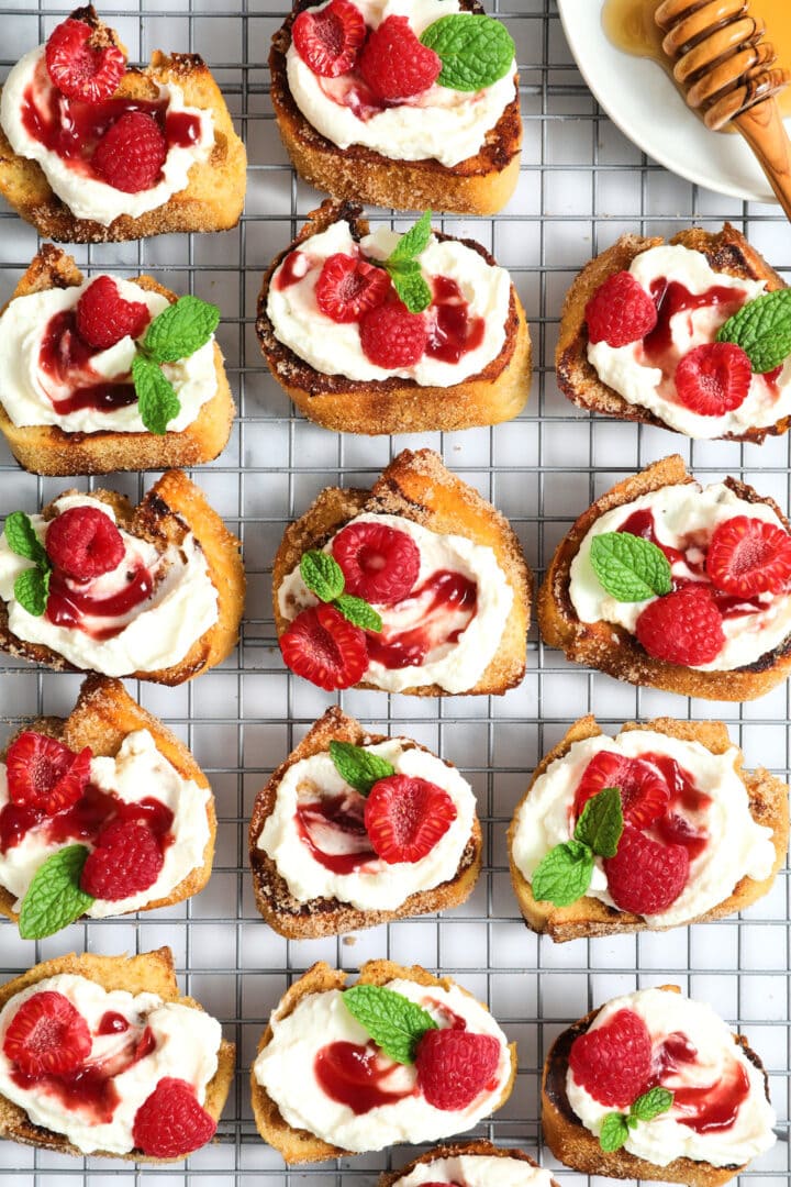 a cooling rack filled with French toast crostini and topped with ricotta, raspberry jam and mint leaves