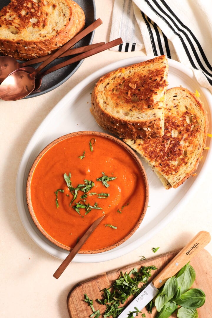 white bowl of tomato soup and two grilled cheese sandwiches on a white plate. There is a small cutting board of chopped basil in the lower right hand corner.