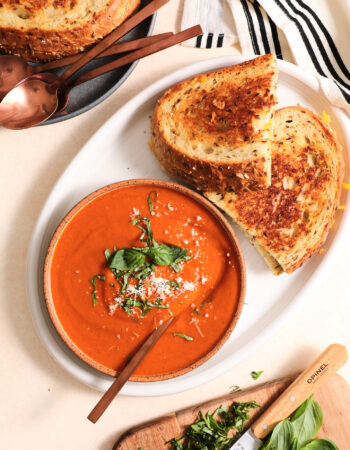 white bowl of tomato soup and two grilled cheese sandwiches on a white plate. There is a small cutting board of chopped basil in the lower right hand corner.