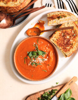 white bowl of tomato soup and two grilled cheese sandwiches on a white plate. There is a small cutting board of chopped basil in the lower right hand corner.