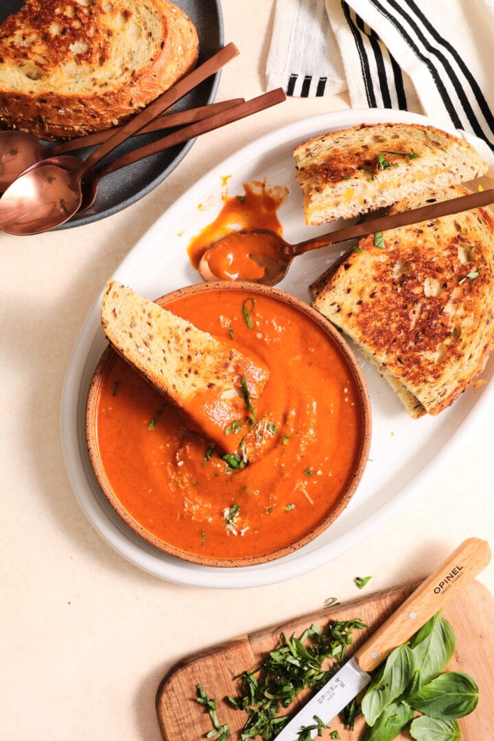 white bowl of tomato soup and two grilled cheese sandwiches on a white plate. There is a small cutting board of chopped basil in the lower right hand corner.