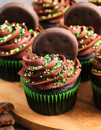 Chocolate cupcakes in green wrappers, with chocolate frosting and green sprinkles. There are small chocolate cookies pressed into the tops of the cupcakes.