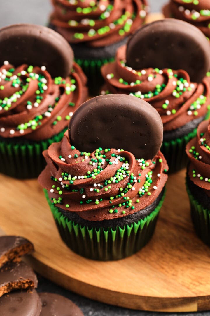 Chocolate cupcakes in green wrappers, with chocolate frosting and green sprinkles. There are small chocolate cookies pressed into the tops of the cupcakes.