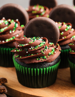 Chocolate cupcakes in green wrappers, with chocolate frosting and green sprinkles. There are small chocolate cookies pressed into the tops of the cupcakes.