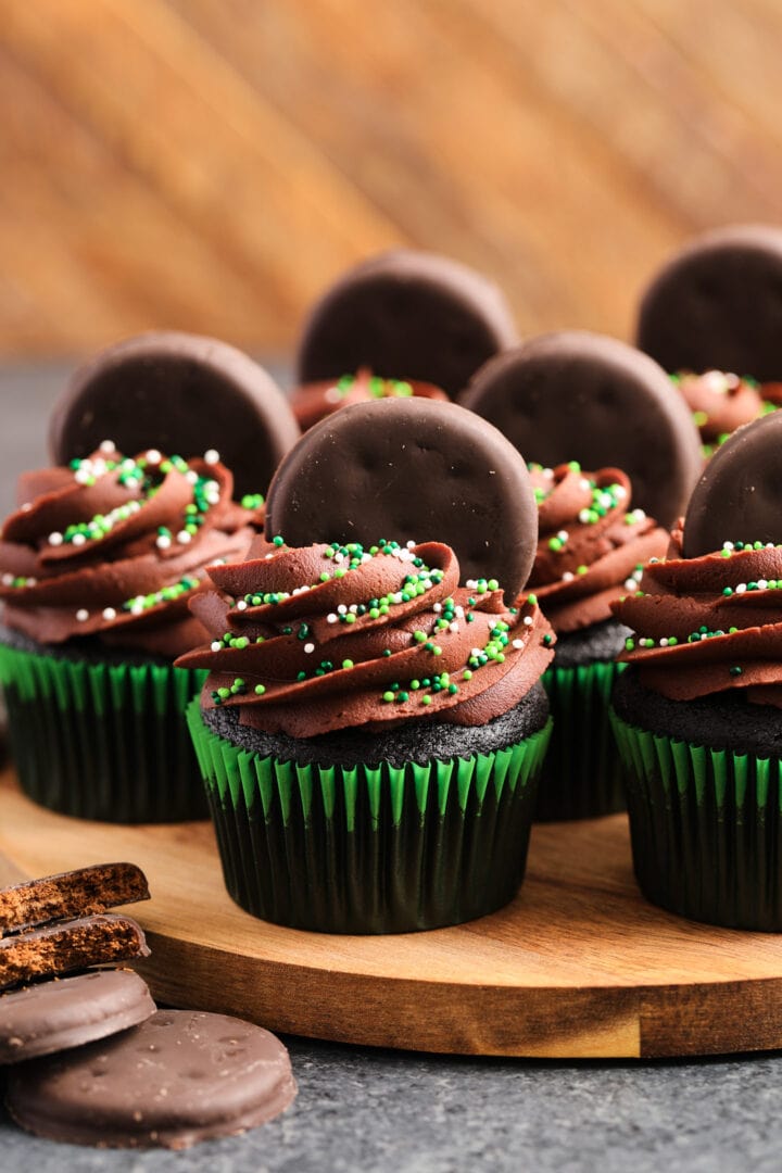 Chocolate cupcakes in green wrappers, with chocolate frosting and green sprinkles. There are small chocolate cookies pressed into the tops of the cupcakes.