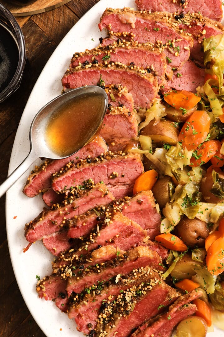 white platter with slices of corned beef brisket and vegetables, including cabbage, carrots and potatoes. There is a silver spoon drizzling broth over the meat.