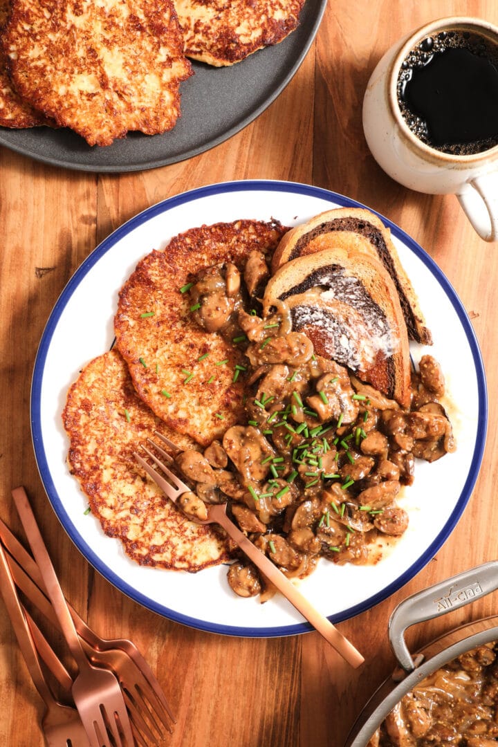 plate of potato pancakes, mushroom gravy and chives on a white plate with a cup of coffee to the right.