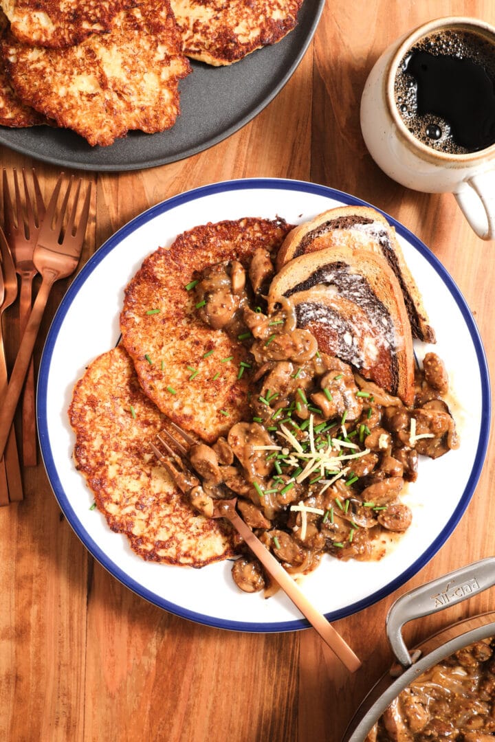 plate of potato pancakes, mushroom gravy and chives on a white plate with a cup of coffee to the right.