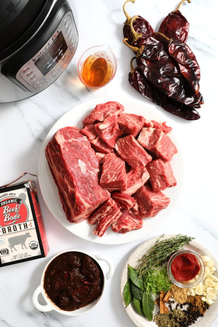 ingredients to make beef birria tacos, including the beef, herbs and spices and peppers. There is an instant pot cooker to the top left.