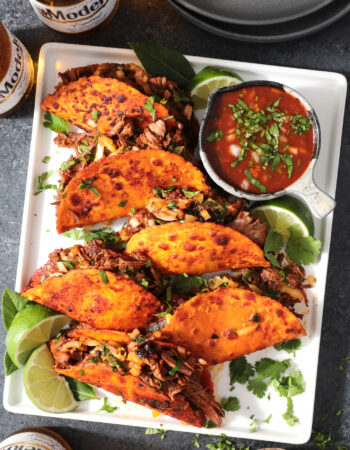 white stray of tacos, garnished with lime, cilantro and a small bowl of red broth to the side.