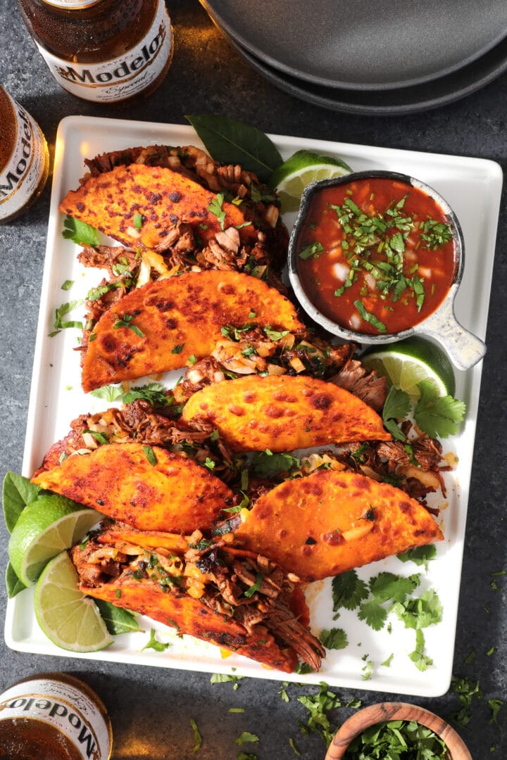 white stray of tacos, garnished with lime, cilantro and a small bowl of red broth to the side.