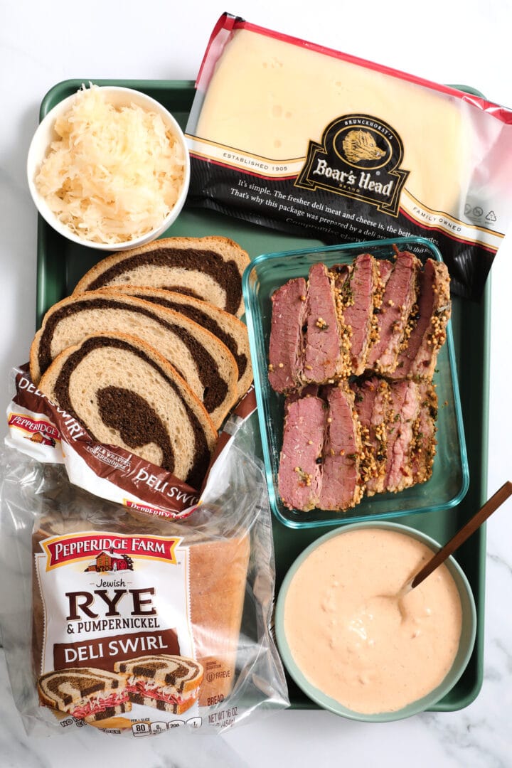 necessary ingredients to make reuben sandwiches, including corned beef, swiss cheese, rye bread, sauerkraut and Russian dressing.