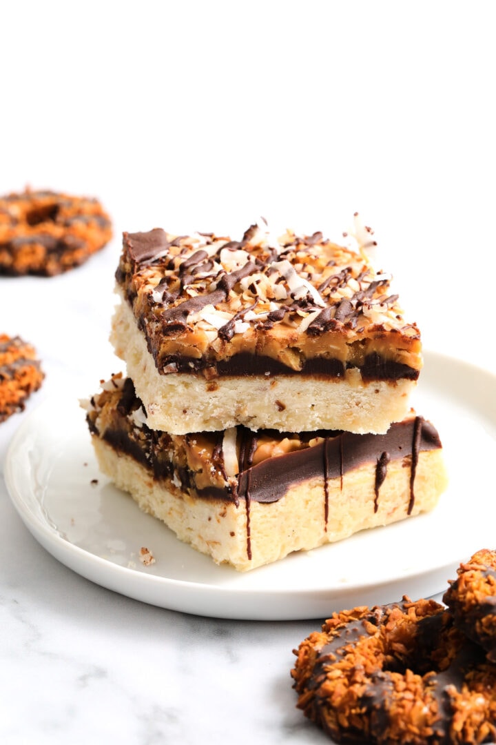 two shortbread cookie bars topped with chocolate, caramel and toasted coconut flakes. There are several other round cookies in the background.