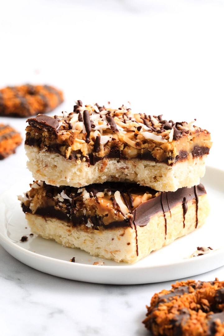 two shortbread cookie bars topped with chocolate, caramel and toasted coconut flakes. There are several other round cookies in the background.