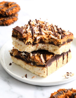 two shortbread cookie bars topped with chocolate, caramel and toasted coconut flakes. There are several other round cookies in the background.