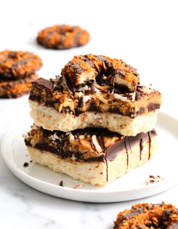 two shortbread cookie bars topped with chocolate, caramel and toasted coconut flakes. There are several other round cookies in the background.