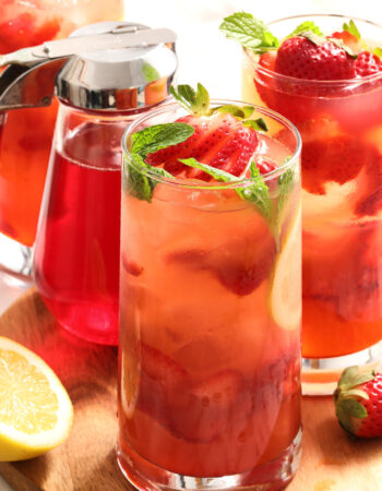 three glasses of strawberry lemonade with mint leaves and a pitcher of strawberry syrup.