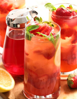 three glasses of strawberry lemonade with mint leaves and a pitcher of strawberry syrup.