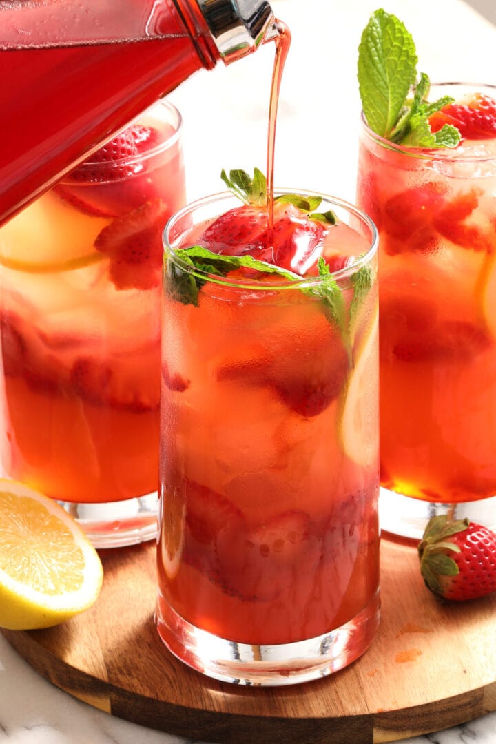 three glasses of strawberry lemonade with mint leaves and a pitcher of strawberry syrup.