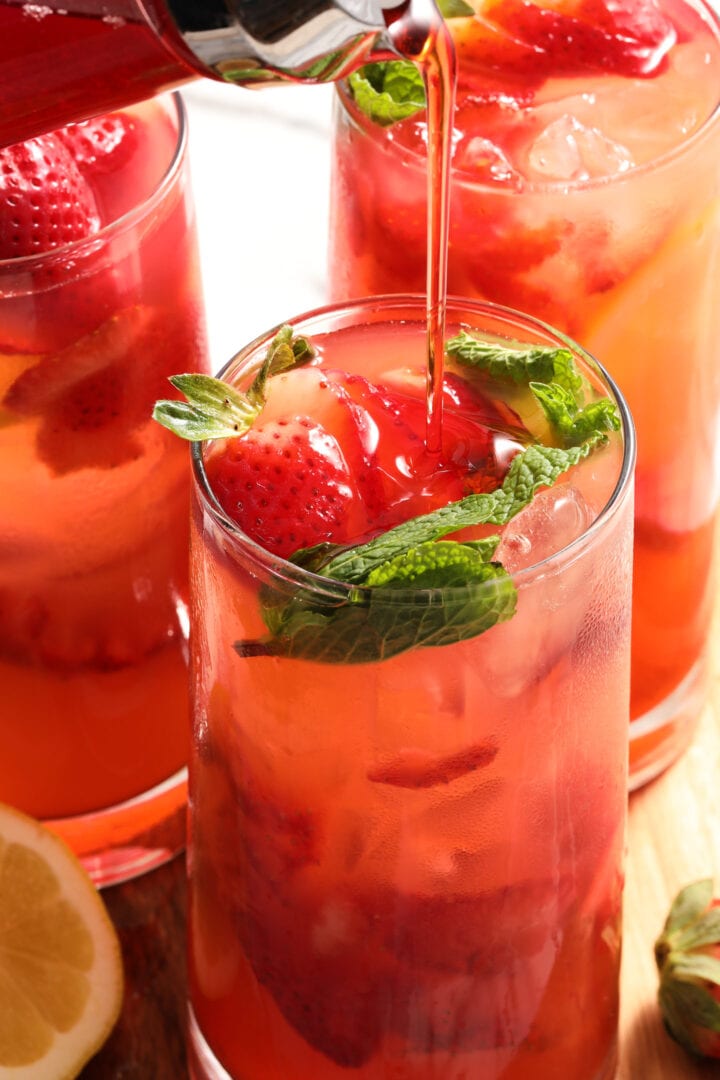 three glasses of strawberry lemonade with mint leaves and a pitcher of strawberry syrup.