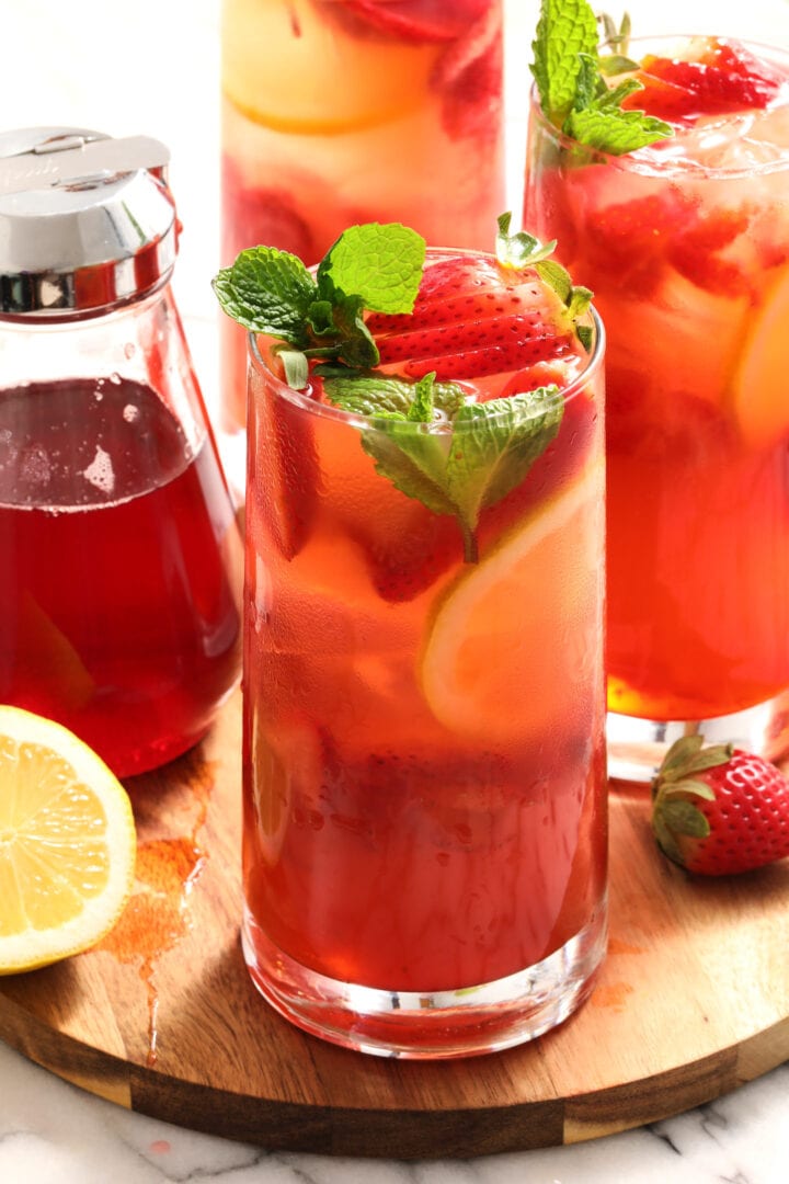 three glasses of strawberry lemonade with mint leaves and a pitcher of strawberry syrup.