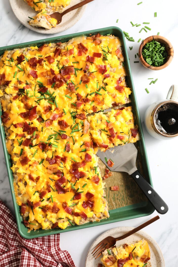 top view of an entire green sheet pan of breakfast pizza, with a few slices on speckled plates off to the side. There is a small wooden bowl of chives and a cup of coffee on the right hand side.