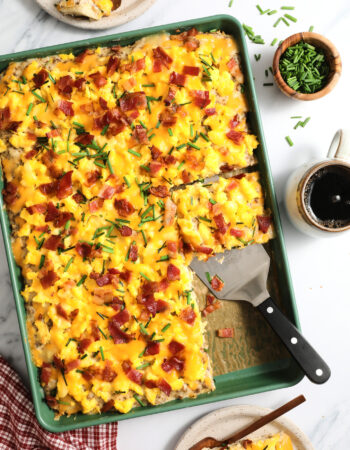 top view of an entire green sheet pan of breakfast pizza, with a few slices on speckled plates off to the side. There is a small wooden bowl of chives and a cup of coffee on the right hand side.