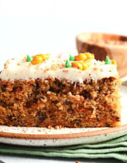 slice of carrot cake with small carrots piped on top of the white frosting. There is a small bowl of pecans in the background.