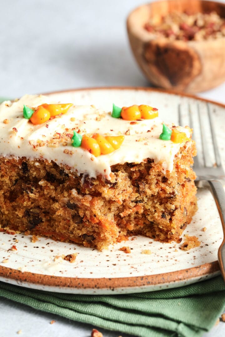 slice of carrot cake with small carrots piped on top of the white frosting. There is a small bowl of pecans in the background.