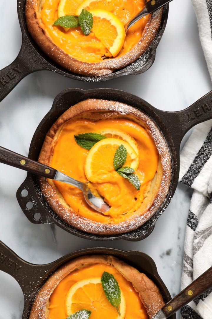 three small cast iron skillets each holding a puffy Dutch pancake, filled with orange curd and garnished with mint leaves and powdered sugar.