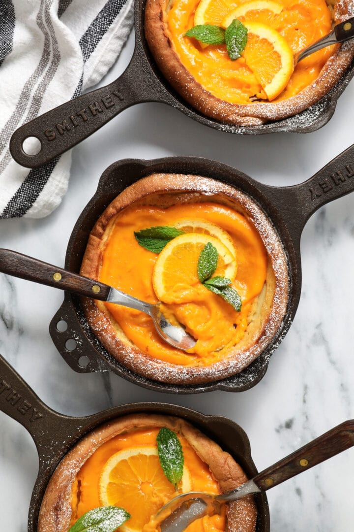 three small cast iron skillets each holding a puffy Dutch pancake, filled with orange curd and garnished with mint leaves and powdered sugar.