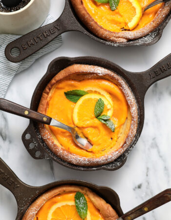 three small cast iron skillets each holding a puffy Dutch pancake, filled with orange curd and garnished with mint leaves and powdered sugar.