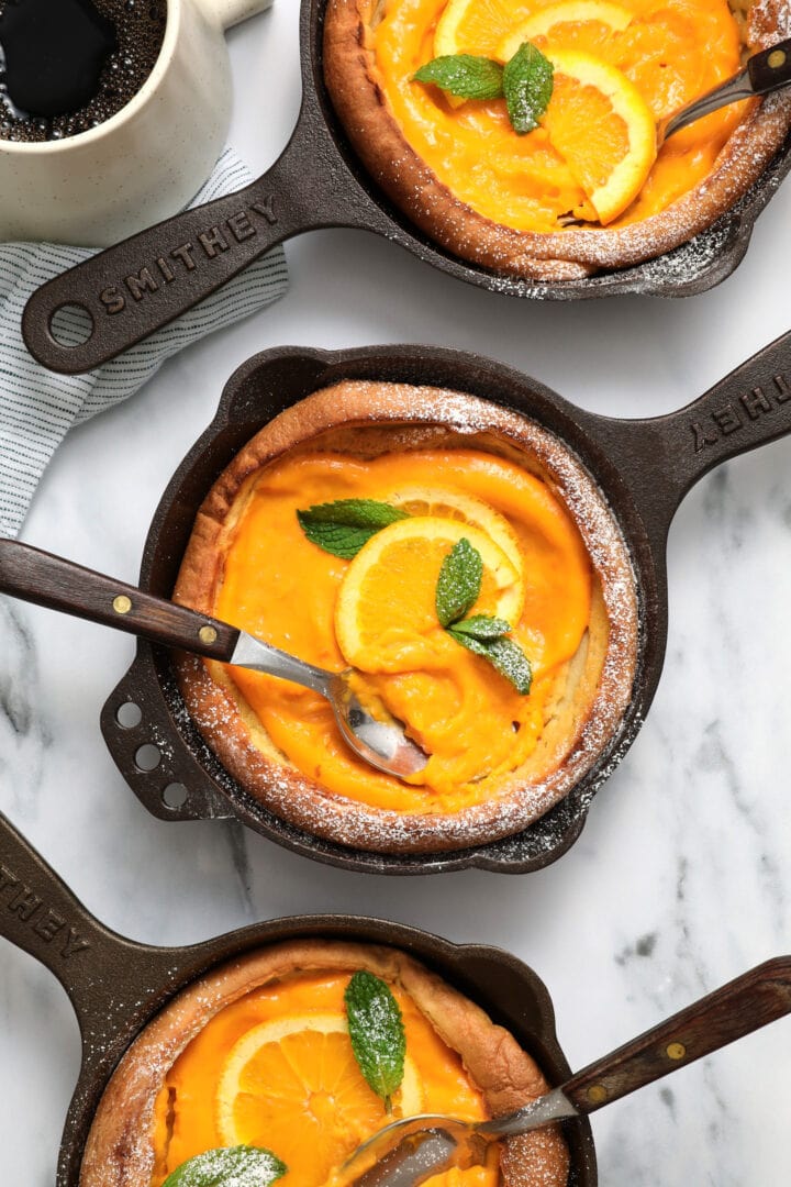 three small cast iron skillets each holding a puffy Dutch pancake, filled with orange curd and garnished with mint leaves and powdered sugar.