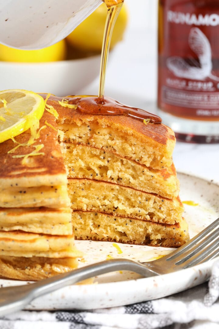 stack of lemon poppyseed pancakes being drizzled with syrup, there are bites missing from the right hand side of the stack.