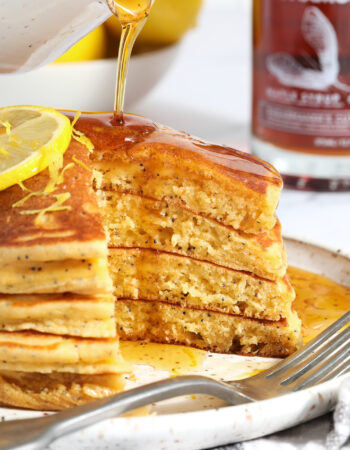 stack of lemon poppyseed pancakes being drizzled with syrup, there are bites missing from the right hand side of the stack.