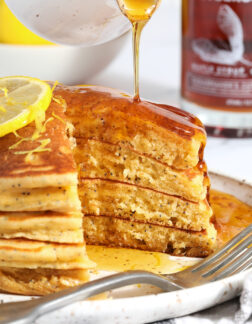 stack of lemon poppyseed pancakes being drizzled with syrup, there are bites missing from the right hand side of the stack.