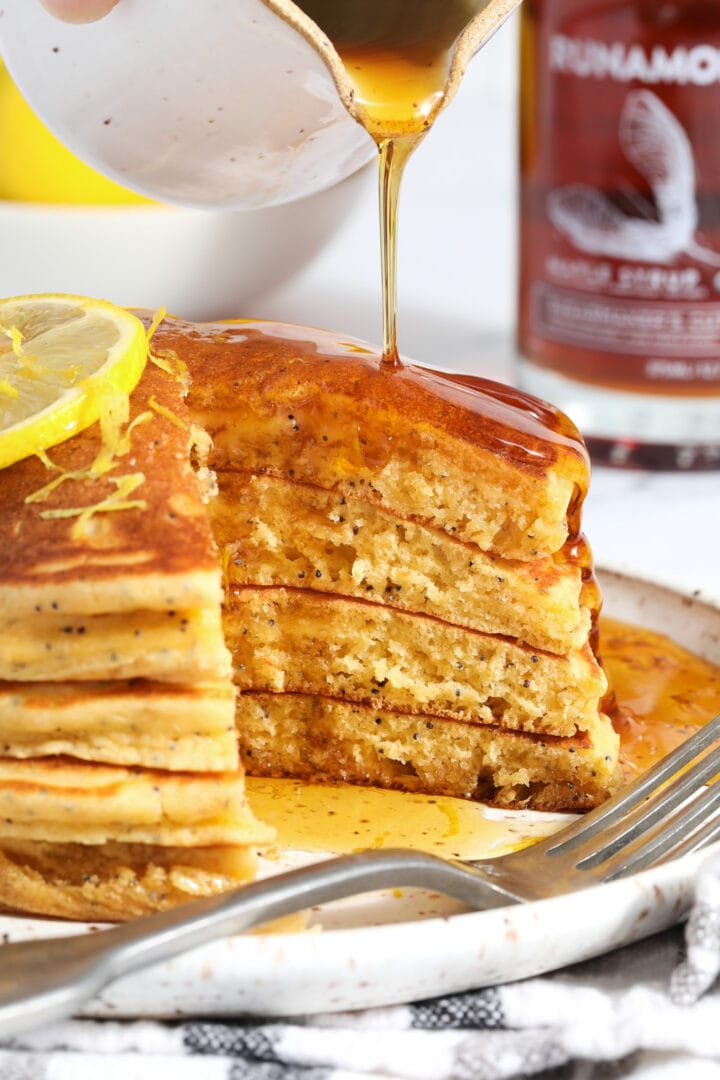 stack of lemon poppyseed pancakes being drizzled with syrup, there are bites missing from the right hand side of the stack.