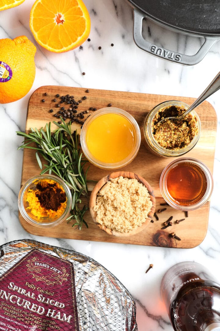 assorted ingredients to make a baked ham, including the ham, orange juice, orange zest, ground cloves, fresh rosemary, peppercorns, brown sugar and mustard.