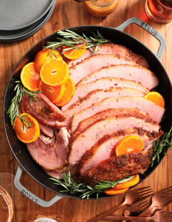 large gray skillet with ham slices arranged with cut oranges and fresh rosemary sprigs.