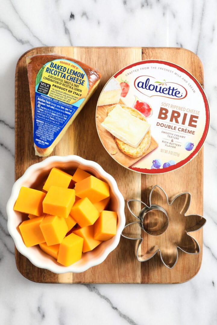 assorted cheeses for the charcuterie board, including a small wheel of brie, wedge of lemon flavored ricotta, and a small bowl of cheddar cubes.