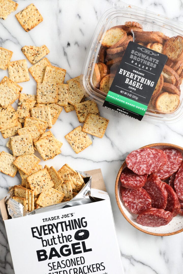 a box of everything bagel seasoned crackers and a small white plate of sliced salami.