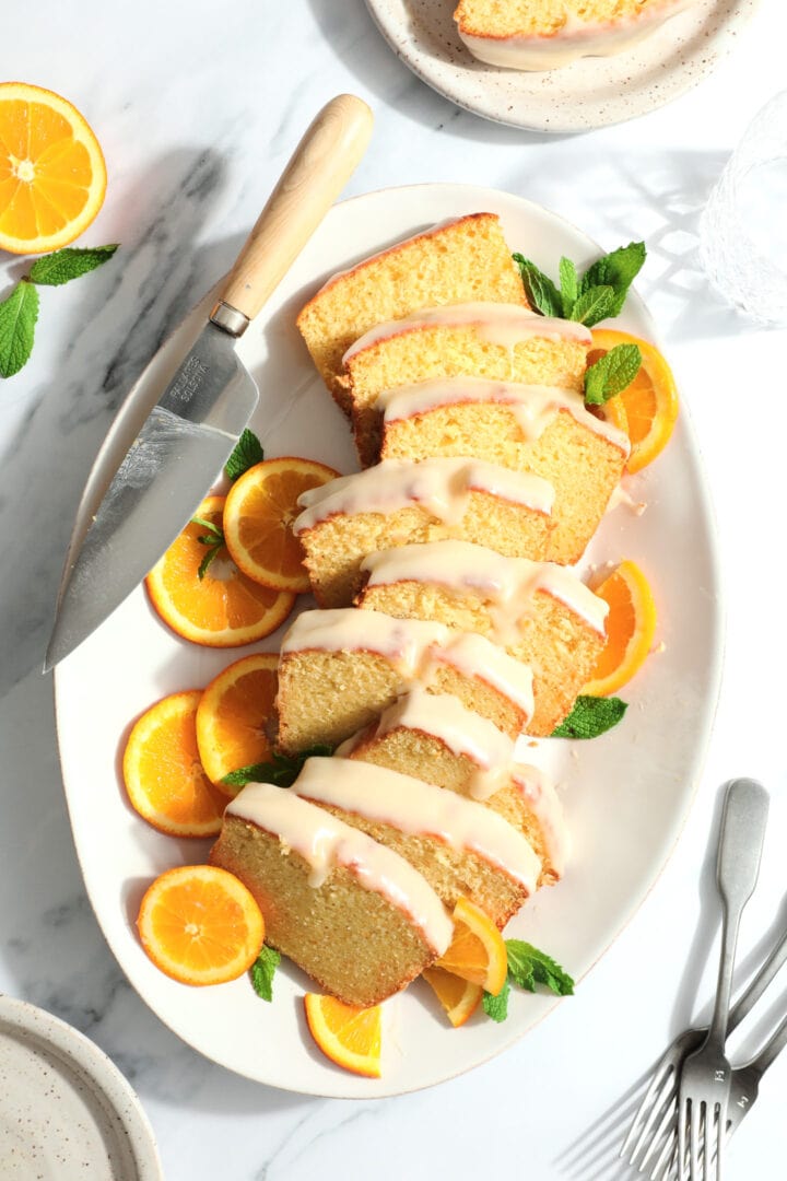 a sliced loaf cake on a white oval platter. The cake slices are glazed with a white frosting and sprinkled with orange zest.