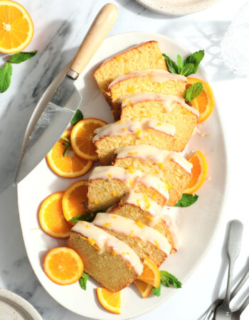 a sliced loaf cake on a white oval platter. The cake slices are glazed with a white frosting and sprinkled with orange zest.